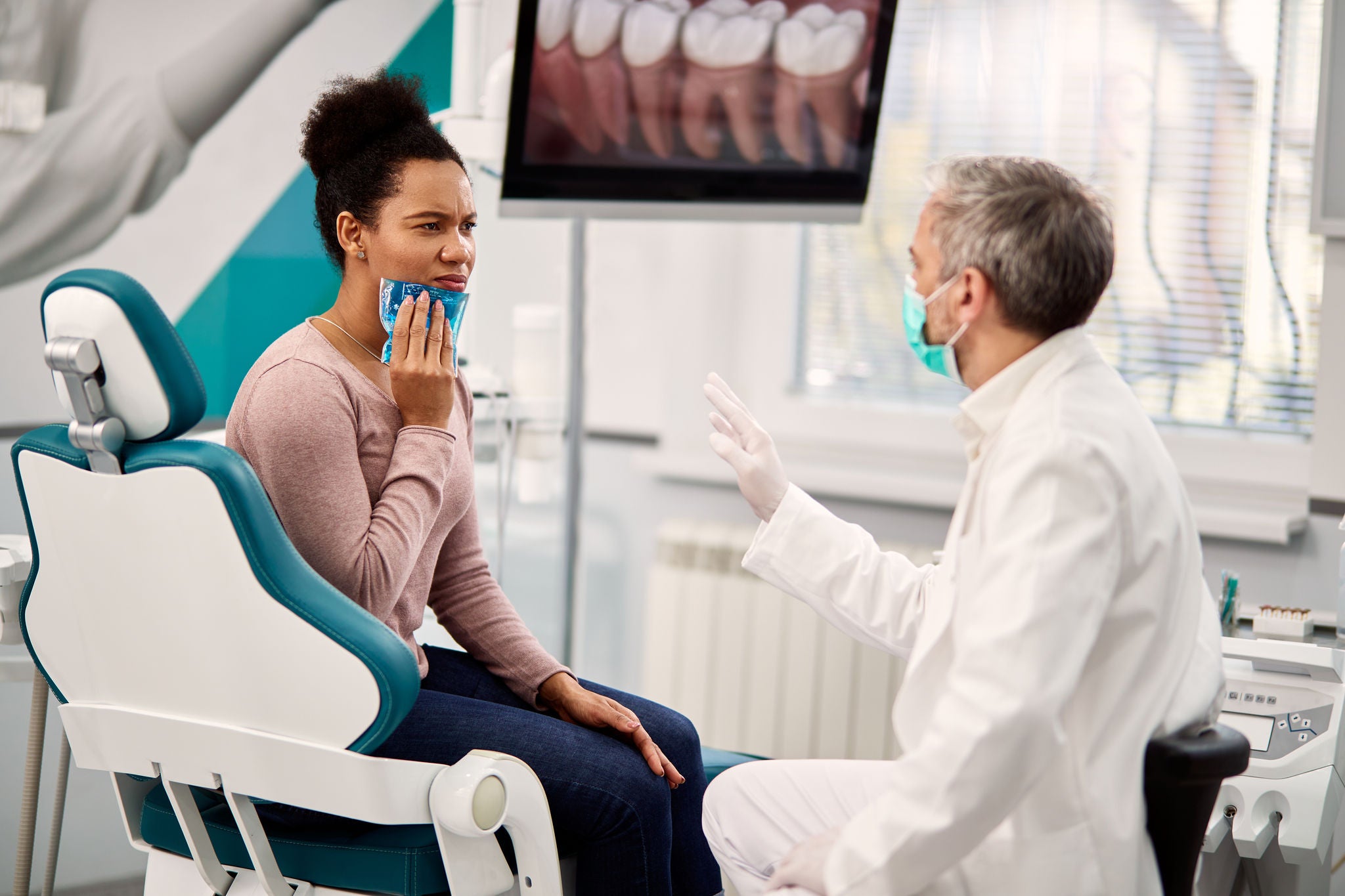 African American woman with toothache holding cooling gel on her jaw while communicating with stomatologist during dental check-up at dentist's office. 