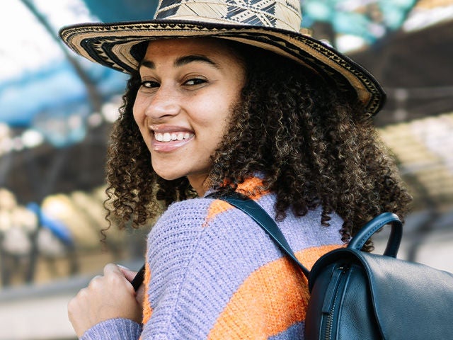 A smiling woman with curly hair wears a wide-brimmed hat and a colorful sweater, carrying a black backpack. Background is blurred. Resolution: 640x480.