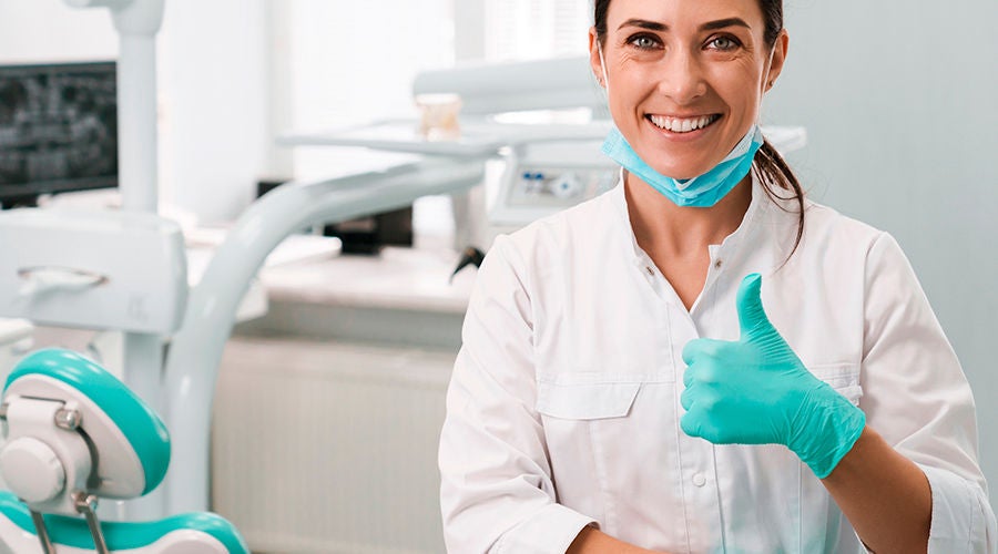 A smiling female dentist in a white coat and teal gloves gives a thumbs-up in a modern dental office. Image resolution: 1920x1080.