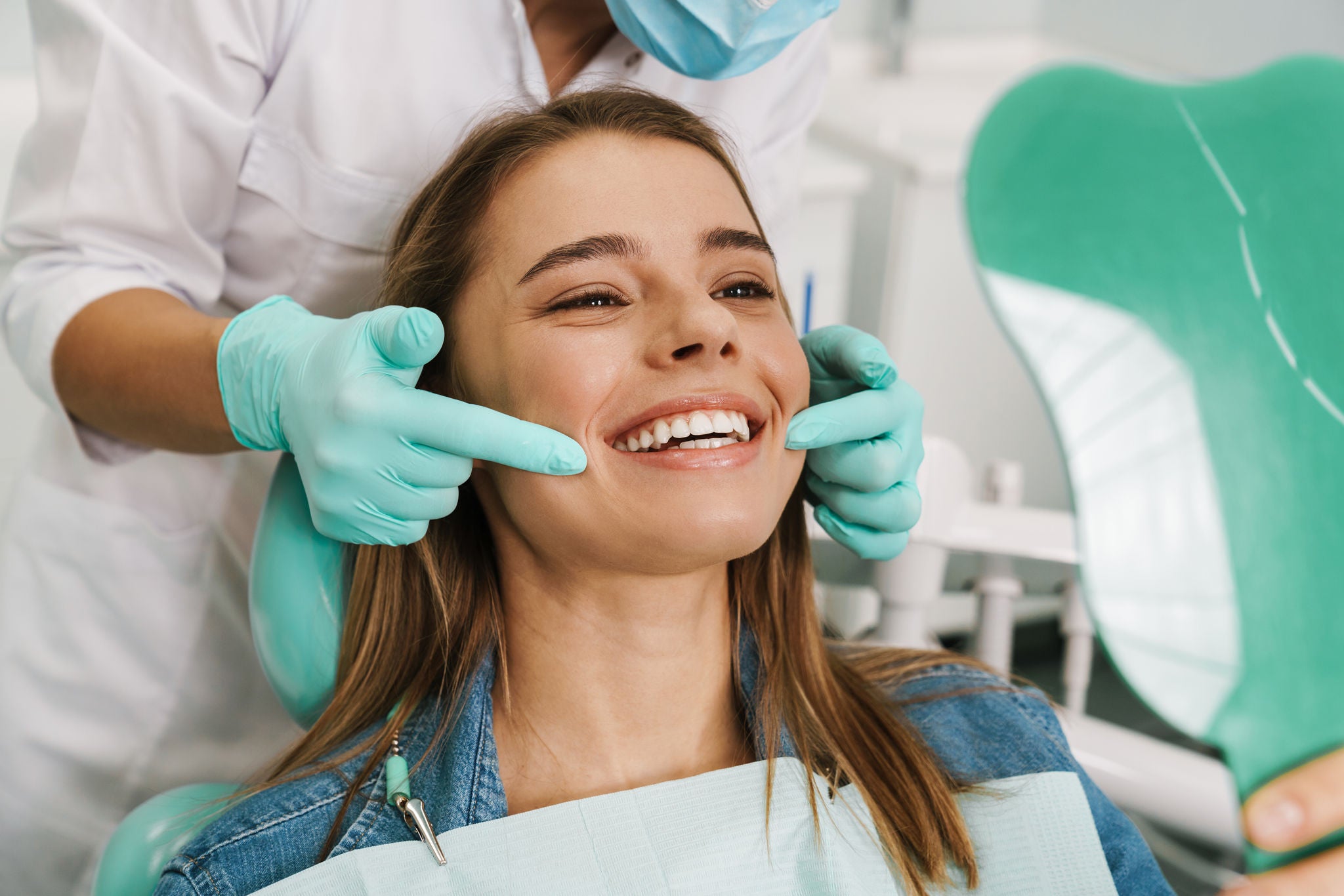 A smiling woman in a dental chair is having her cheeks gently examined by a dentist wearing gloves and a mask. Image resolution: 1920x1280.